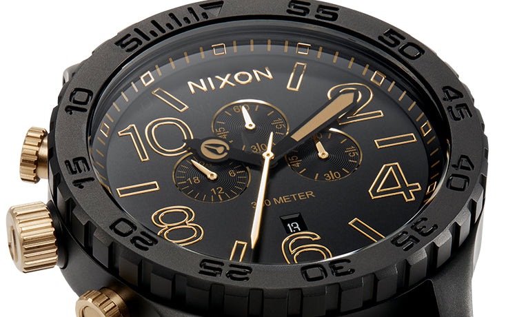 Close-up of a Nixon 5130 Chrono watch with a matte black case, gold numbers, hands, and sub-dials on a black face, and gold crown and pushers.