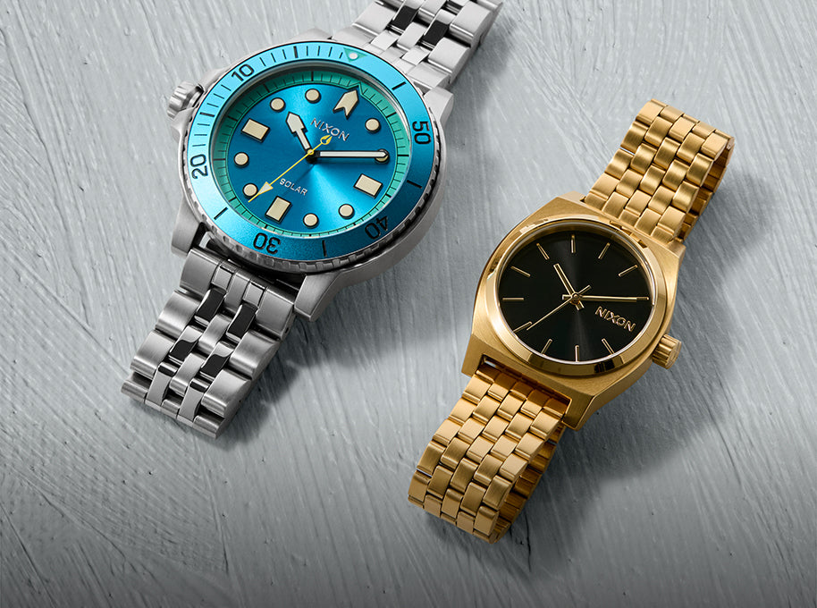 Two Nixon watches: a silver dive watch with a blue face and a gold watch with a black face.