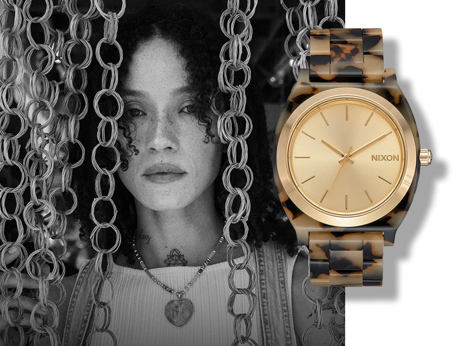 Black and white portrait of a woman with curly hair and freckles, partially obscured by chains, next to a gold Nixon watch with a tortoiseshell band.