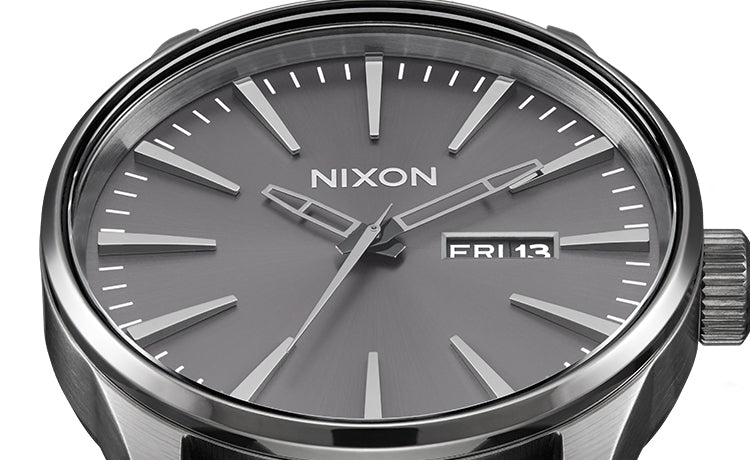 Close-up of a Nixon Sentry watch with a gunmetal grey dial, silver hour markers, and "FRI 13" date display.