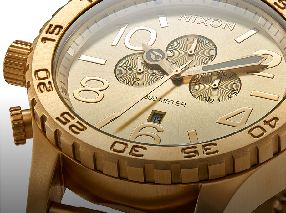 A close-up of a gold Nixon chronograph watch face with three sub-dials, large numerals, and a date window.