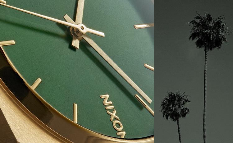 Close-up of a Nixon watch with a green dial and gold accents, next to two silhouetted palm trees against a dark sky.